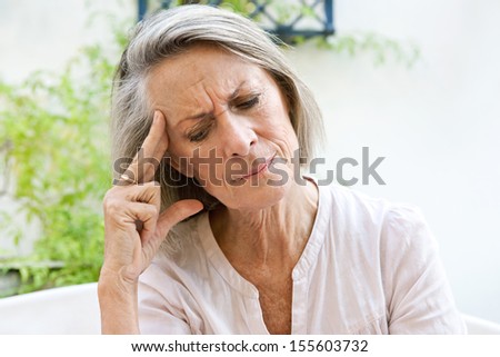 Mature woman sitting on a white sofa in a home garden touching her head with her hand and fingers while having a headache pain and feeling unwell, outdoors.