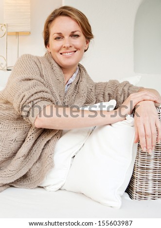 Portrait of an attractive middle aged woman laying and relaxing on a white sofa in a neutral living room at home, smiling at the camera and being serene, interior.
