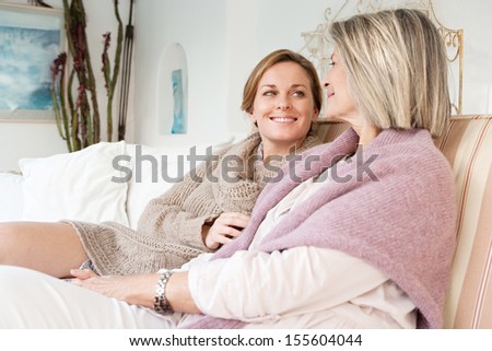 Attractive And Alike Adult Daughter And Mature Mother Relaxing Together And Being Affectionate With Each Other While Sitting On A White Sofa At Home And Smiling, Interior.