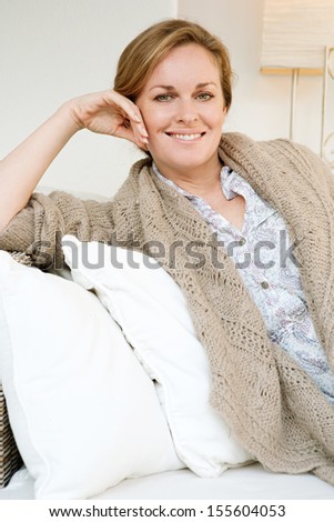 Portrait of an attractive and confident middle aged woman sitting and relaxing on a white sofa in a neutral living room at home, smiling at camera and being serene, interior.