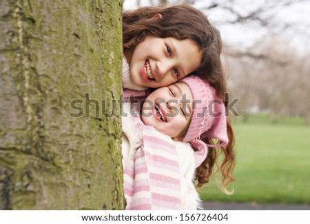 Close up portrait of two young child girl sisters family with their heads together peeking and standing next to a tree trunk in a park during a cold winter day playing games, outdoors.