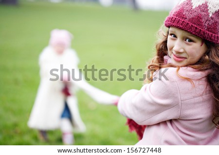 Two young sisters children playing tag of war against each other, pulling a woolly scarf and competing in a green park field during a cold winter day, outdoors.