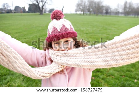 Portrait view of a joyful and welcoming young girl being playful and opening her arms in a park during a cold winter day, wearing a scarf and smiling while having fun, outdoors.