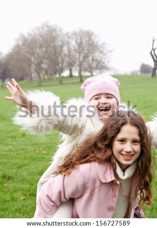 Older sister giving younger sister a piggy bag while playing and having fun in a green park grass field during a cold winter day outdoors, smiling joyful.
