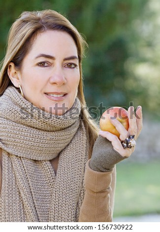 Portrait of an attractive hispanic middle aged mature woman in a home garden holding a red apple fruit in her hand, looking at camera with space, outdoors.