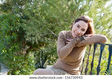 Portrait of an attractive hispanic middle aged mature woman in a home garden relaxing and leaning on an iron balcony banister, looking at camera and smiling with space, outdoors.