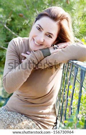 Portrait of an attractive hispanic middle aged mature woman in a home garden relaxing and leaning on an iron balcony banister, looking at camera and smiling, close up.