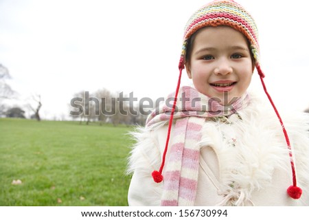 Portrait of a young child girl wearing a coat and a rainbow woolly hat and scarf during a cold winter day in a green park field with a cloudy sky, smiling joyfully outdoors.