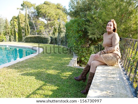 Proud home owner relaxing in her country home garden with trees and a swimming pool during a sunny autumn morning, sitting on a stone wall with veranda balcony.