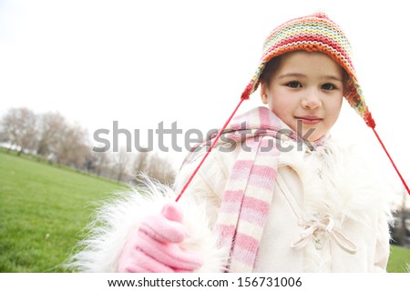 Close up of a young child girl wearing a coat and a rainbow woolly hat during a cold winter day in a green park, smiling softly and pulling the knitted hat strings, outdoors.