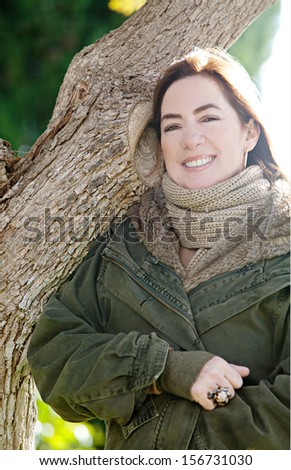 Close up portrait of an attractive middle aged mature woman visiting the countryside and leaning on an autumn tree trunk during a sunny morning, wearing a coat and smiling.