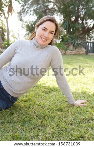 Portrait of an attractive hispanic middle aged mature woman relaxing on the green grass of a home garden during a sunny fall day, joyful and smiling outdoors.