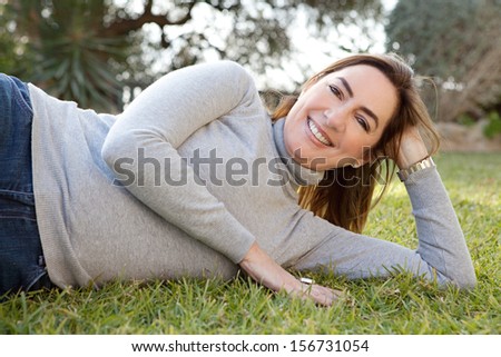 Portrait of an attractive hispanic middle aged mature woman in a home garden relaxing and laying down on green grass during a sunny autumn day, outdoors.