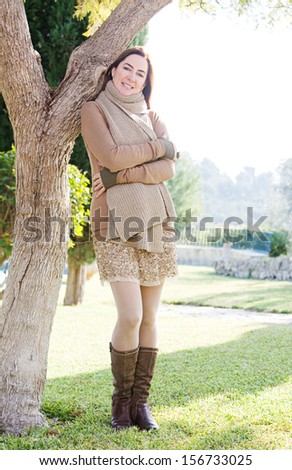 Attractive middle aged mature woman visiting a rural home garden and leaning on an autumn tree trunk during a sunny morning, enjoying the garden and wearing a scarf and boots.
