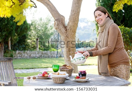 Side view of an attractive mature woman pouring and serving tea with a tea pot while enjoying a healthy breakfast in a home garden during an autumn morning, outdoors.
