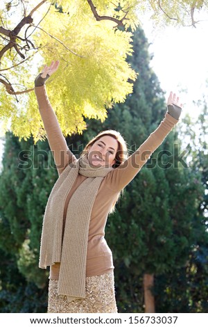 Attractive mature woman reaching with her arms outstretched up to the changing leaves of a tree while visiting a park during an autumn sunny morning, outdoors.