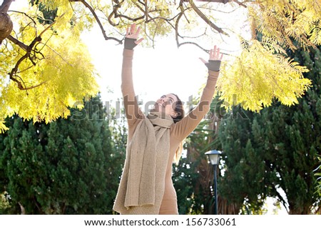 Attractive mature woman reaching with her arms outstretched up to the changing leaves of a tree while visiting a park during an autumn sunny morning, outdoors.