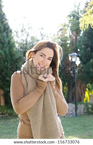 Close up portrait of an attractive middle aged smiling woman in a park, holding her woolly knitted scarf around her neck keeping warm during a sunny winter morning, outdoors.