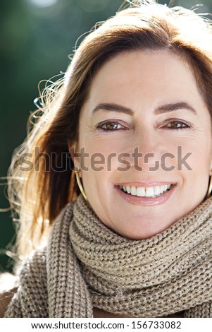 Close up portrait of an attractive joyful middle aged woman in a park, holding her hair up and smiling, keeping warm during a sunny winter morning, outdoors.