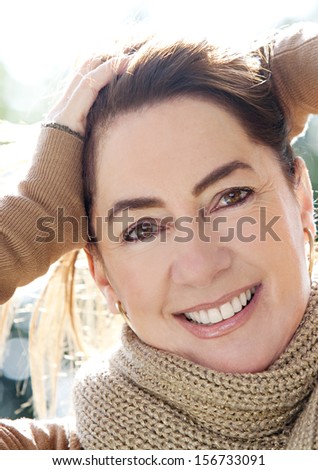 Close up portrait of an attractive joyful middle aged woman in a park, holding her hair up and smiling, keeping warm during a sunny winter morning, outdoors.