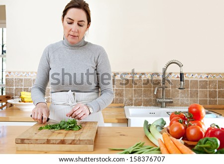 Attractive mature woman chopping herbs and cooking vegetables in the kitchen at home, with healthy organic produce and using a knife and chopping board.