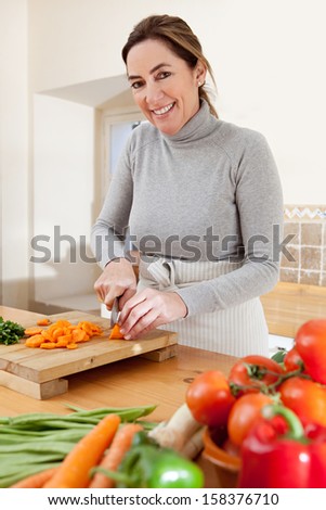 Attractive mature woman chopping herbs and carrots, and cooking vegetables in the kitchen at home, with healthy organic produce and smiling at the camera, indoors.