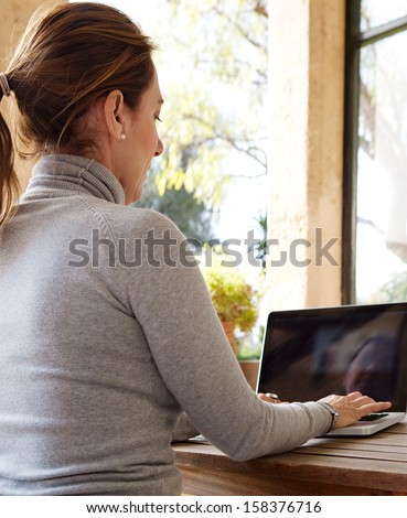 Rear side view of a smart professional woman typing on her laptop computer while sitting near a large glass window at home overlooking the garden during a sunny day.