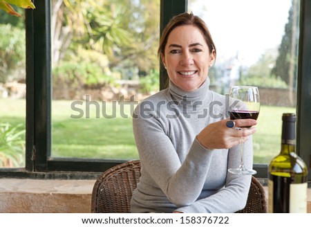 Portrait of an attractive mature woman sitting in a home interior with large glass windows and a green garden, relaxing drinking wine and smiling at the camera, indoors.