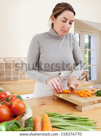 Attractive mature woman chopping herbs and carrots, and cooking vegetables in the kitchen at home, with healthy organic produce and using a knife and a wooden chopping board.