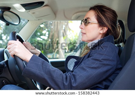 Side portrait view of a mature middle aged smart woman sitting on the drivers seat of a new car, driving through a countryside road during a sunny winter day.