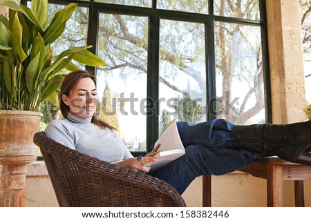 Attractive mature woman with her feet up on a table, sitting in a home conservatory with large glass windows and a green garden, reading a book and relaxing indoors, smiling.