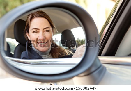 Reflection in a car reversing side mirror of a smart and mature woman driver, smiling at the camera while driving and arriving at home, feeling proud and happy.