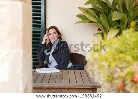 Portrait of a mature woman relaxing at home with a book, smiling at the camera while sitting near green plants and leaves, feeling positive and confident.