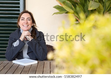 Portrait of a joyful mature woman relaxing at home with a book, leaning on her hands and smiling at the camera while sitting by a wooden table, feeling positive and confident.