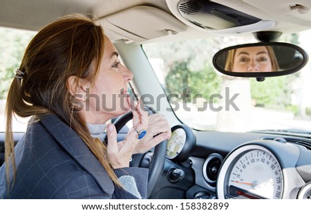 Side portrait view of a mature middle aged smart woman sitting on the drivers seat of a new car, retouching her make up cosmetics using the rear mirror while parked.