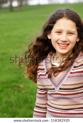 Portrait of a joyful and active young girl child with long red curly hair, running in a green grass field in a park during an autumn sunny day, wearing a stripy jumper outdoors.