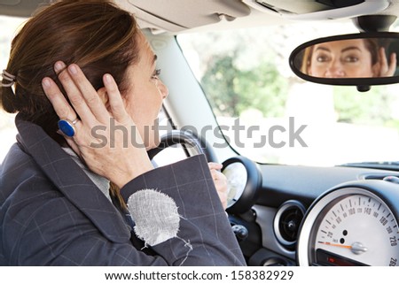 Side portrait view of a mature middle aged smart woman sitting on the drivers seat of a new car, retouching her hair using the rear mirror reflection while parked.