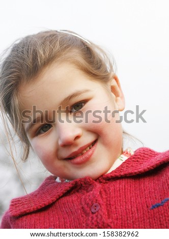 Close up portrait of a beautiful young girl with a gentle smiling expression, wearing a red knitted jumper against the sky while in a park during winter autumn day outdoors.