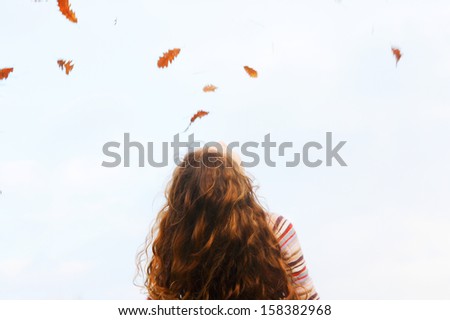 Spacious rear view of a child girl with red curly hair tilting her head back and looking up to the sky with dry autumn leaves floating and falling down, outdoors.