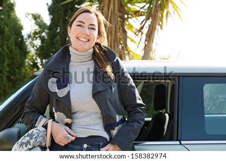 Smart and elegant mature middle aged woman leaning on her new transport car auto during a sunny winter day, feeling proud and smiling at the camera joyfully.