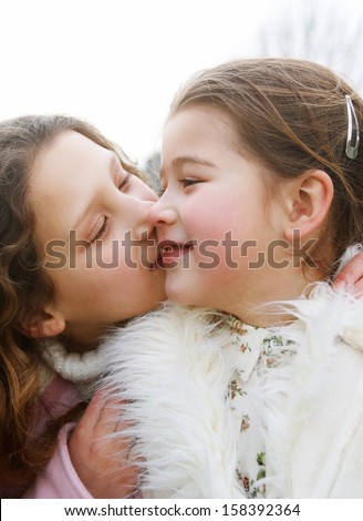 Close up portrait of two young girls children sisters together in a park during a cold winter day, with one kissing the other on the cheek, smiling and enjoying the outdoors.
