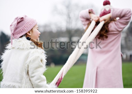Two young joyful sisters family children playing together and having fun with games, pulling a woolly scarf in a green park field during a cold winter day, outdoors.