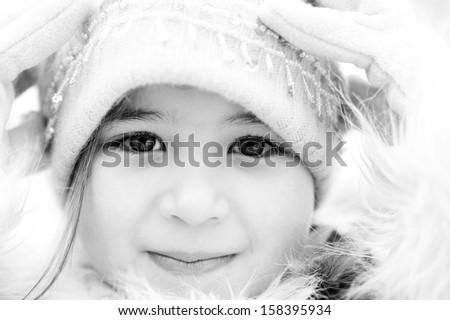 Black and white close up beauty portrait of a young child girl wearing a winter coat, woolly hat and gloves, smiling to the camera during a cold winter day, outdoors.