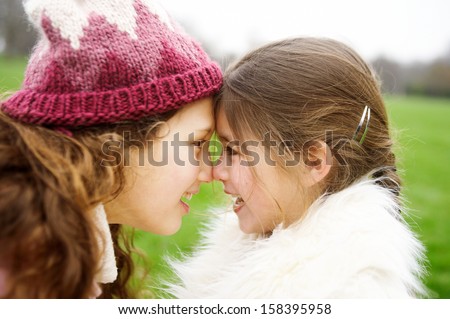 Close up profile portrait of two girl children sisters rubbing noses together while in a park during a cold winter day, having fun and smiling outdoors.