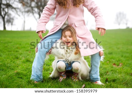 Two young children and family sisters playing together in a park with green grass and bare trees during a cold winter day, wearing coats and having fun outdoors.