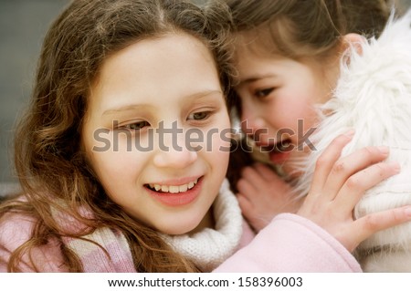 Close up portrait of two young girls children sisters in a park during a cold winter day, with one whispering secrets into the others ear, smiling and laughing outdoors.