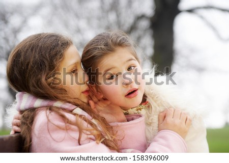 Close up portrait of two young girls children sisters sitting on a wooden bench in a park during a cold winter day, with one whispering secrets into the others ear, smiling outdoors.