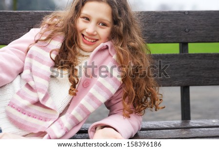 Beautiful young girl with red curly hair relaxing and laying down on a wooden bench in a park during a cold winter day, wearing a pink coat and a scarf, smiling outdoors.