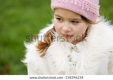Close up portrait of a beautiful young child girl standing in a green park looking away and being thoughtful during a cold winter day, wearing a warm coat and a pink hat, outdoors.