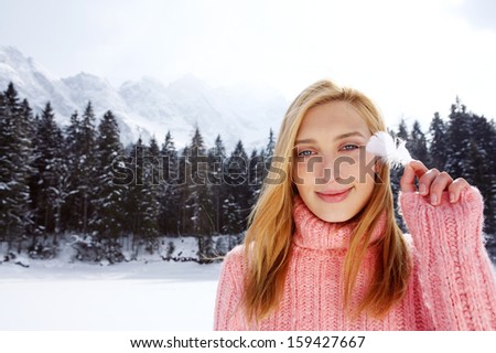 Close up portrait of a beautiful young woman on vacation in the snow mountains, caressing her face with a natural white feather and smiling with tickles during a sunny winter day, outdoors.